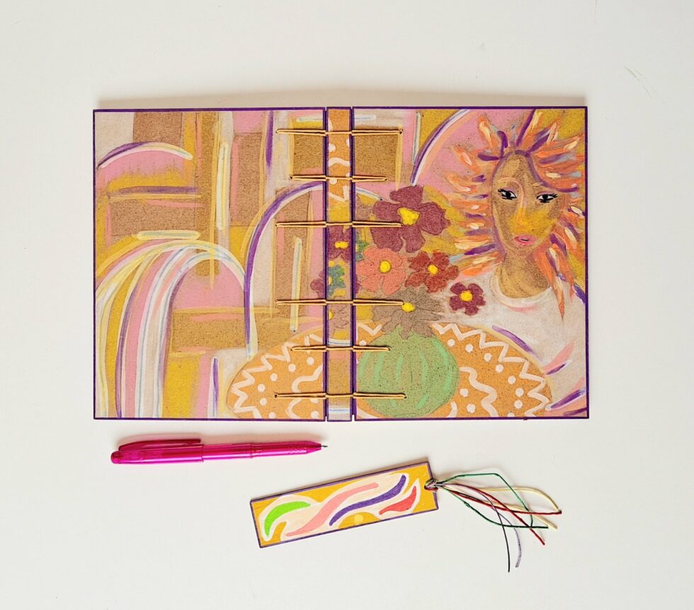 Carnet SUZANA posé ouvert avec un stylo rose et un marque-page en bois à côté.