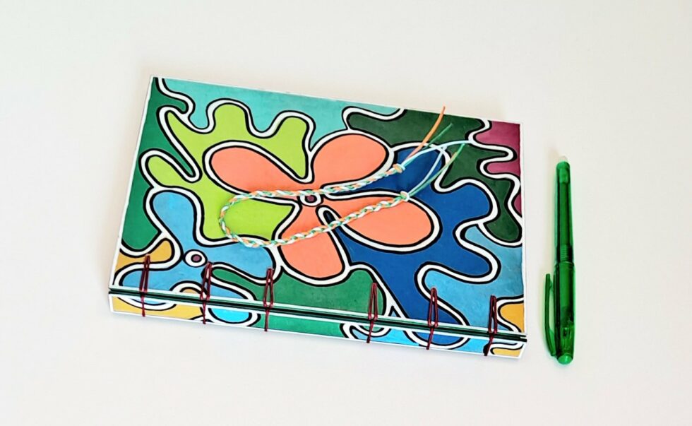 Carnet CAPUCINE, vue recto à plat dos avec reliure fil rouge et stylo vert.