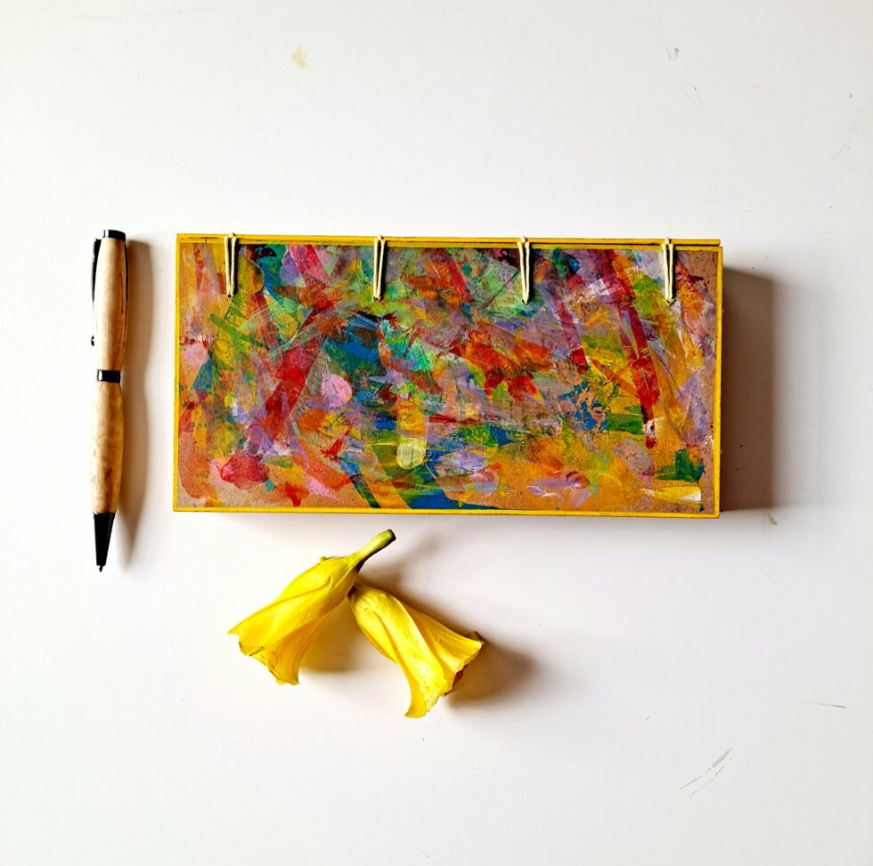 Boîte à mots OASIS vue recto fermée sur reliure jaune avec stylo en bois et 2 fleurs jaunes.