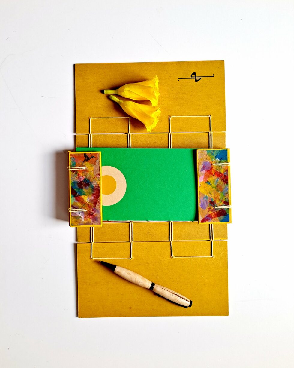 Boîte à mots OASIS vue recto 2 côtés ouverts sur reliure jaune avec stylo en bois et 2 fleurs jaunes.