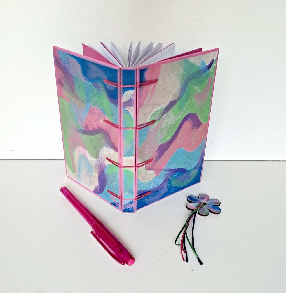 Carnet PASTEL vue recto verso debout reliure rose avec marque pages en bois peint et stylo rose.