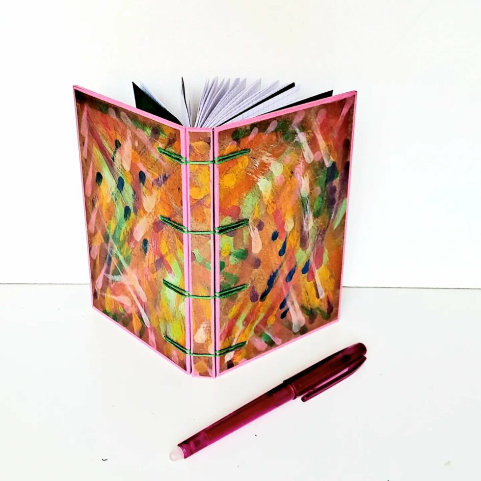 Carnet MONA, vue recto verso debout avec marque-pages en bois et stylo rose.