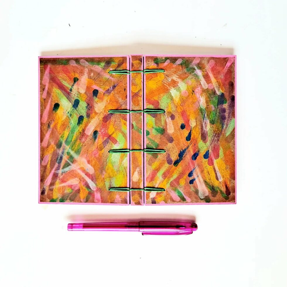 Carnet MONA, vue recto verso à plat avec marque-pages en bois et stylo rose.