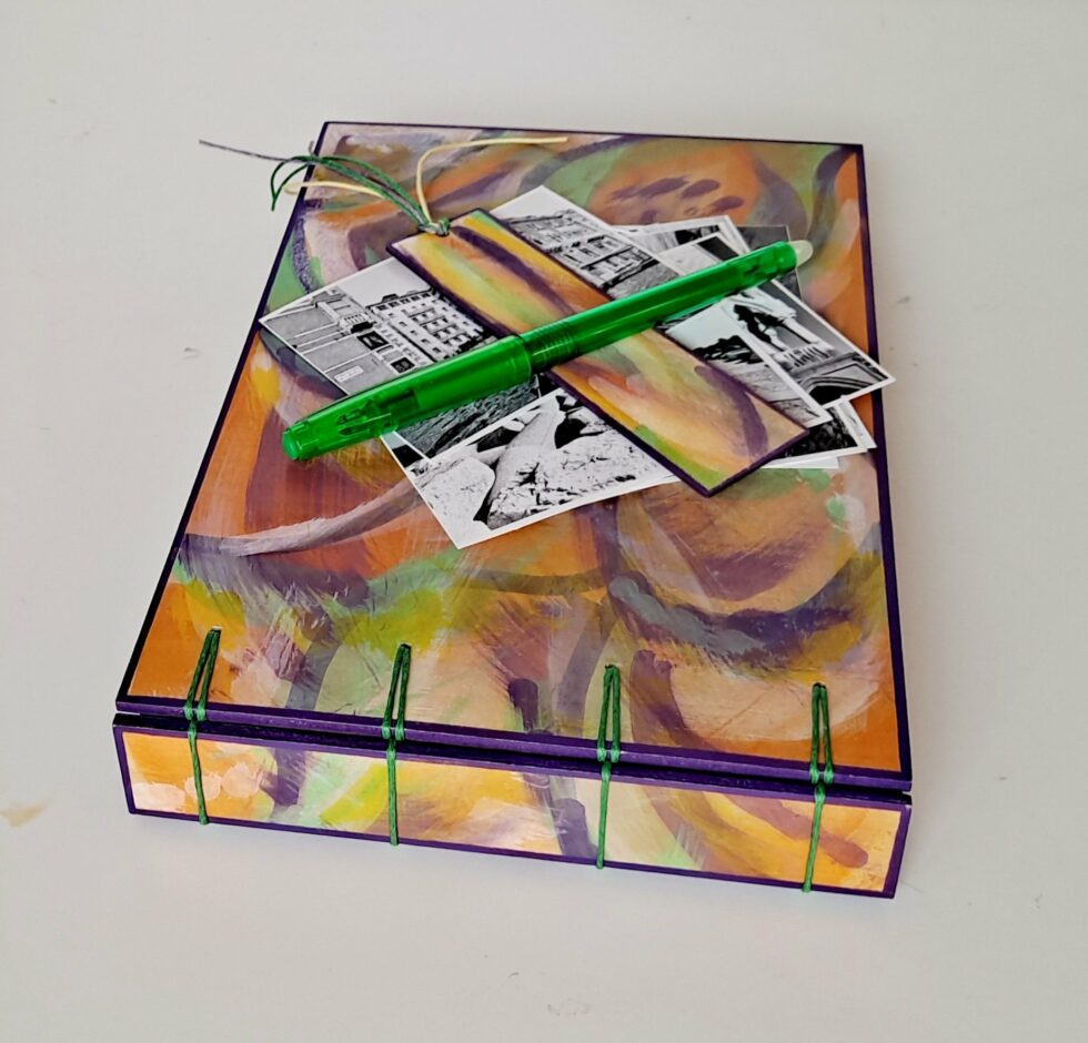 Album MAHÉ, vue recto à plat sur dos avec marque-pages en bois peint et stylo vert.