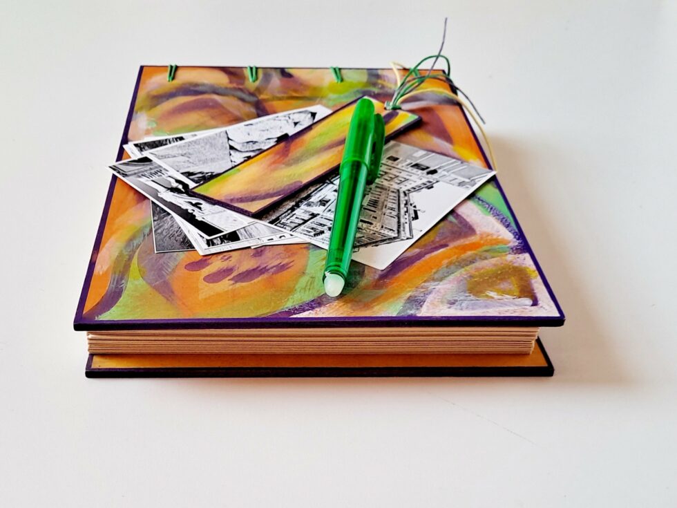 Album MAHÉ, vue recto à plat sur pages avec marque-pages en bois peint et stylo vert.