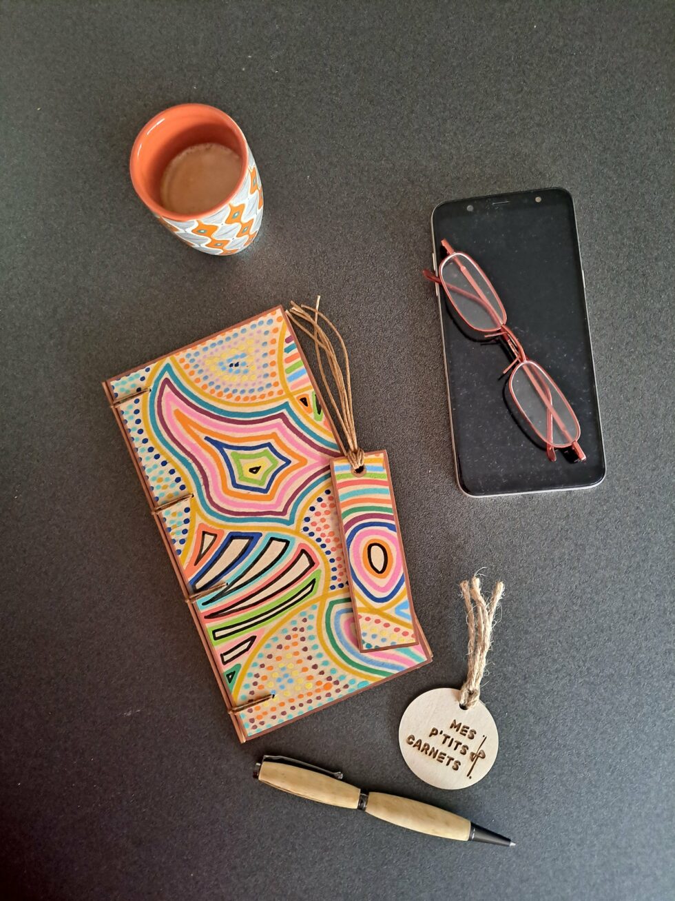 Carnet KIMBA, vue recto avec marque-pages en bois peint à la main et stylo et téléphone et lunettes.