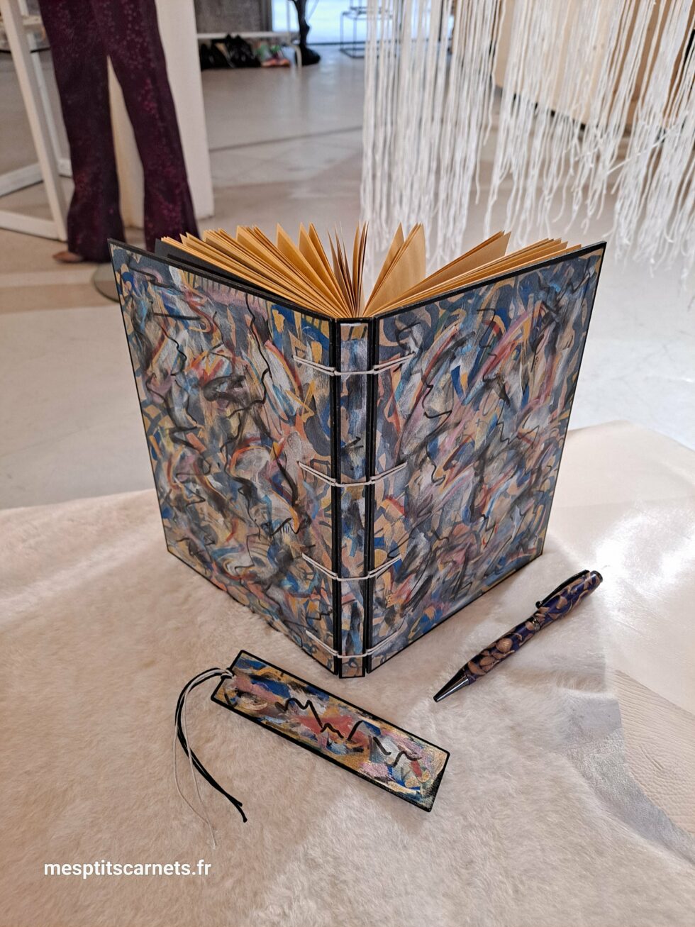 Carnet FRAC, vue recto verso debout avec marque-pages en bois peint et stylo.