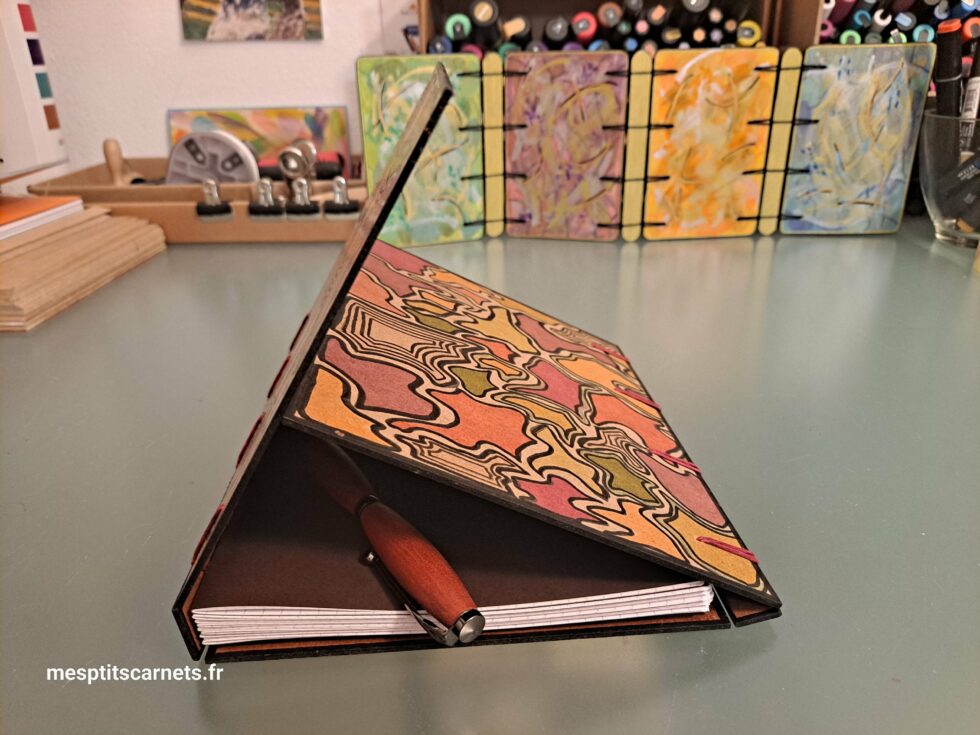 Carnet Triptyque SILICE vue semi ouvert à plat et couverture en bois peint et stylo.